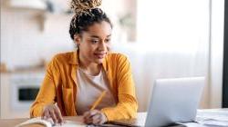 Young black woman with dreadlocks sitting at desk with laptop open and writing on pen and paper 